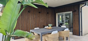 Patio with WoodUpp vertical slatted wood cladding and black trim profiles covering the corner wall, a grey rectangular outdoor dining table, six woven rattan chairs with white cushions, a small potted centerpiece, sliding glass door, and large tropical leaves framing the foreground.