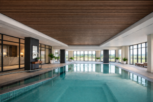 A luxurious indoor swimming pool area with large windows overlooking a natural landscape. The high ceiling is covered with WoodUpp's dark wood acoustic slat panels, featuring narrow wooden strips that run across the length of the room, complementing the modern design.