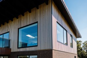 WoodUpp vertical slatted timber cladding covers the upper storey, a natural pale wood profile forming narrow, evenly spaced vertical battens around corners and window openings. WoodUpp soffit panels under the deep eaves complement large black-framed glazing above a brick base, creating a modern, textured façade.