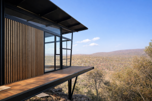 Exterior view of a modern cliffside structure featuring WoodUpp acoustic panels on the facade. These panels display warm, oak-finish wooden slats against a dark background, complementing the adjacent wooden deck and large glass doors framed in black steel overlooking a vast landscape.