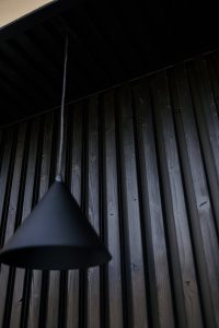WoodUpp image of vertically grooved dark wooden cladding panels, showcasing natural grain and recessed fixing screws, paired with a hanging matte black cone pendant lamp suspended by a slim cable. The minimalist composition emphasizes WoodUpp's timber wall system and complementary contemporary lighting.