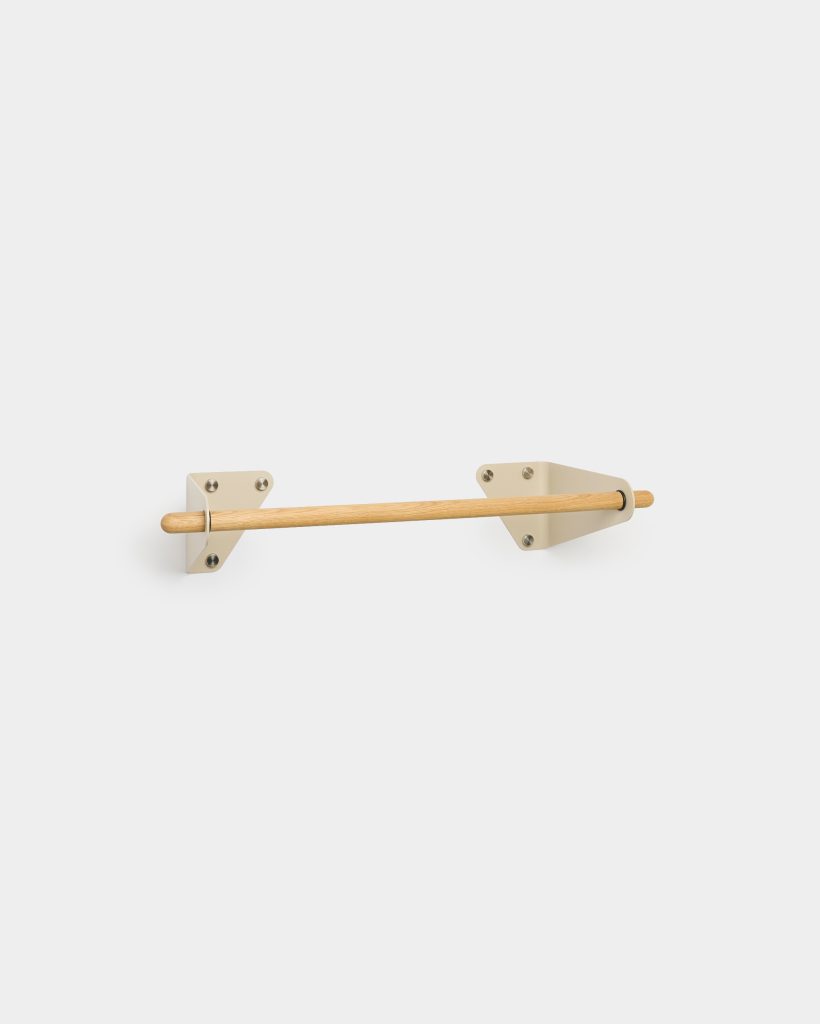 WoodUpp wall-mounted oak dowel rail held by two cream powder-coated metal brackets, shown horizontally against a white wall. The minimalist setup features a smooth cylindrical light-wood bar seated in triangular bracket cups with visible fixing screws, suitable as a towel rail, clothes rod or display fixture.