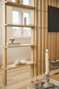 WoodUpp light oak room divider features a freestanding shelving unit and matching vertical slat panel, both with slim metal fixings and round metal feet. Shelves hold books, yarn balls and a silver bowl; the slatted panel supports a wall-mounted TV beside a lit candle in the foreground.