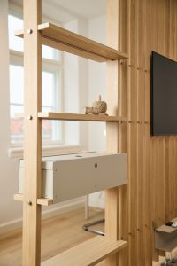 WoodUpp oak ladder-shelf and slatted room divider in light natural finish, shown with a beige WoodUpp metal cable-management box fitted onto a mid shelf. Natural twine spool and small wooden peg rest on the upper shelf, highlighting the unit’s clean, modular storage and modern living-room styling.