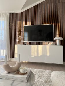 Sunlit living room with WoodUpp vertical slat wall panels in dark walnut forming a textured backdrop behind a wall-mounted TV. Below sits a white floating cabinet topped with a glass vase with twisted branches, stacked books, and a white mushroom lamp; cozy rug, marble coffee table and candles in foreground.