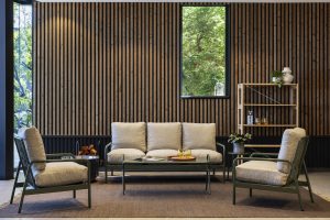 Modern lounge with beige-cushioned metal-frame sofa and two armchairs centered on a brown rug, glass coffee table with glasses, and a tall window. WoodUpp vertical wooden slat wall panels cover the back wall and a WoodUpp freestanding slatted shelving unit on the right holds plants and glassware.